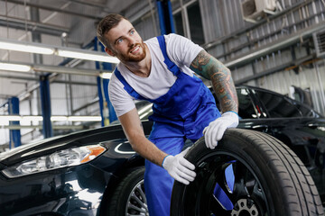 Fototapeta na wymiar Strong minded happy young male professional technician car mechanic man 20s wear denim blue overalls white t-shirt stand hold wheel look aside work in light modern vehicle repair shop workshop indoor