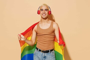 Young blond latin gay man 20s with make up in beige tank shirt headphones wrapped in rainbow flag listen to music dance isolated on plain light ocher background studio People lgbt lifestyle concept.