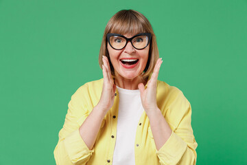 Elderly smiling happy caucasian woman 50s in glasses yellow shirt scream hot news about sales discount with hand near mouth isolated on plain green background studio portrait People lifestyle concept