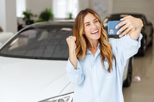 Woman customer buyer in blue shirt do selfie shot mobile phone do winner gesture choose auto want to buy new automobile in car showroom vehicle salon dealership store motor show indoor. Sales concept.