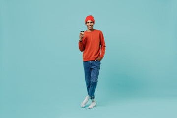 Full body young smiling happy african american man 20s wear orange shirt hat hold takeaway delivery craft paper brown cup coffee to go isolated on plain pastel light blue background studio portrait