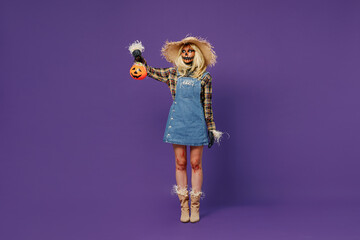 Full body young woman with Halloween makeup mask wearing straw hat scarecrow costume hold look at Jack-o'-lantern pumpkin isolated on plain dark purple background studio Celebration holiday concept.