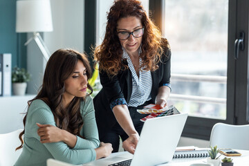 Two beautiful designer women working in a design project with laptop while talking in the office.
