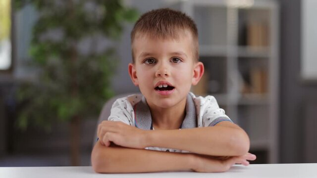 Front view of schoolboy in white polo t-shirt sitting at table and talking on camera. Portrait of cute caucasian child staying at home.