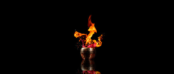 Pote dourado de cerâmica saindo fogo e um liquido vermelho de dentro em um fundo preto. 