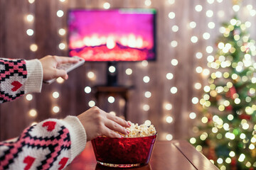 Christmas eve. Alone woman eating popcorn, watching tv, switching channels using remote control. Home cinema. Cropped, close up