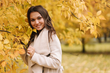 A beautiful pretty young woman in a coat drinks hot coffee walks in the autumn park smiles communicates online and talks using a mobile phone in nature in the fall forest, selective focus