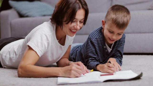 Loving caucasian mother lying on floor with little son and drawing in white album with colorful pencils. Family weekend and children creativity concept.