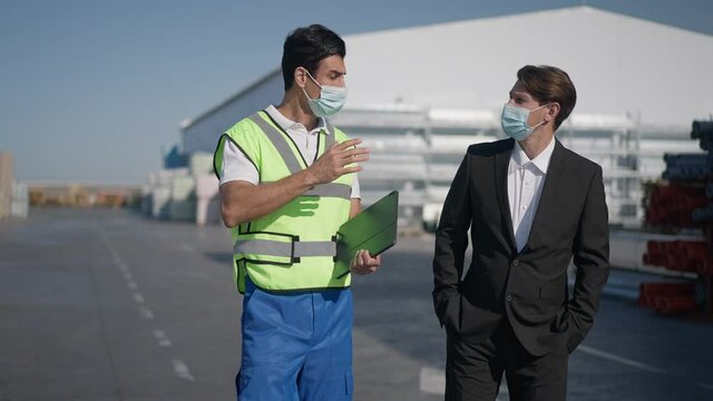 Serious Middle Eastern warehouse worker in uniform and Caucasian supervisor in suit walking talking outdoors on coronavirus pandemic. Portrait of professional men in Covid face masks discussing plan