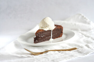 A piece of brownie on a saucer on a light table. On top of the cake is a scoop of ice cream. There is a white towel next to it.