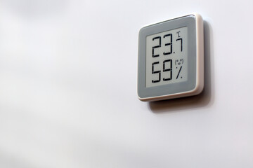 Electronic indoor gray plastic digital battery powered thermometer on white wall showing temperature, humidity level in house, smiling smiley face indoors in daylight sun