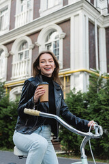 Fototapeta na wymiar cheerful woman holding paper cup while riding bicycle on urban street of europe