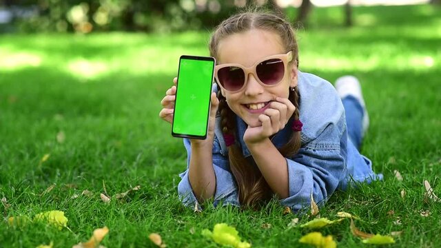 The child is holding a phone with a green screen in nature. A girl lies in the outumn on the grass in a blue dress and looks at the phone