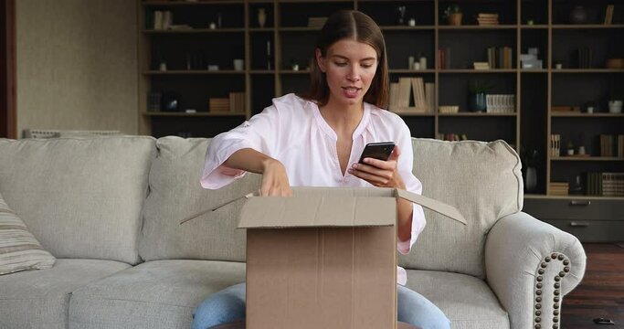 Glad teenage female video blogger sit on couch open parcel with internet shop order shoot unpacking on smartphone front camera. Young lady receive consumer goods from web store record unboxing on cell