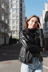 cheerful young woman in leather jacket standing on urban street