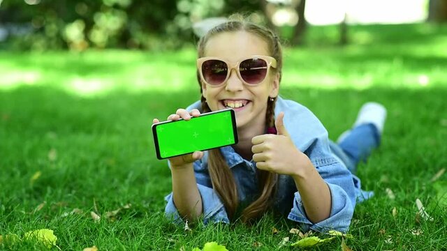 The child is holding a phone with a green screen in nature. A girl lies in the outumn on the grass in a blue dress and looks at the phone