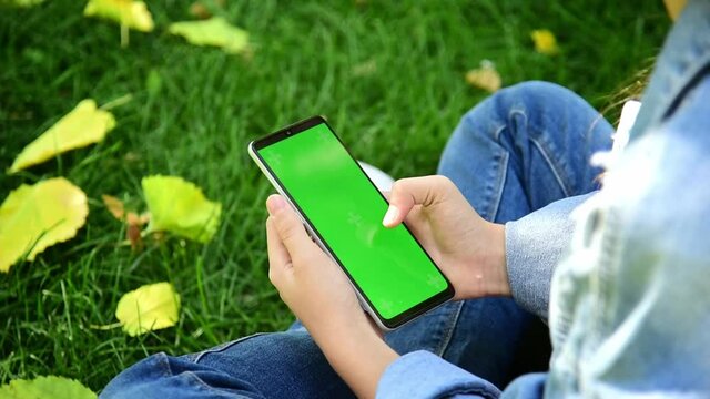 The child is holding a phone with a green screen in nature. A girl lies in the outumn on the grass in a blue dress and looks at the phone