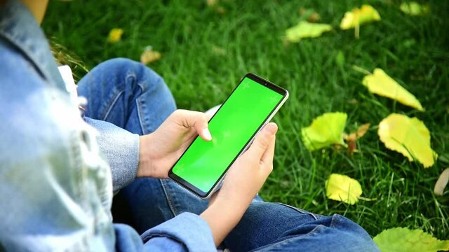 The child is holding a phone with a green screen in nature. A girl lies in the outumn on the grass in a blue dress and looks at the phone