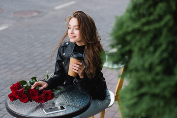 high angle view of pleased woman in leather jacket holding paper cup near smartphone and bouquet of red roses on coffee table - Powered by Adobe