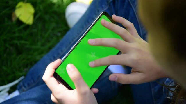 The child is holding a phone with a green screen in nature. A girl lies in the outumn on the grass in a blue dress and looks at the phone
