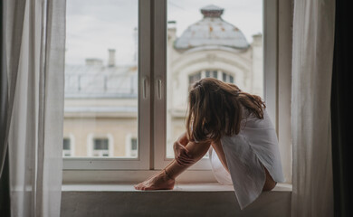 Young sad woman sitting on the windowsill by the window in the apartment hugging her knees and bowing her head. Depression and burnout concept