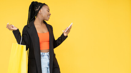 Online shopping. Sale banner. Black Friday. Cheerful Afro woman with shopper bag paying order on phone isolated on orange copy space background.