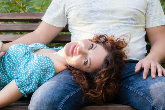 Attractive young woman with red hair lies on the lap of a man on a bench in a city park. Loving couple on a romantic date in the park. Family summer walk. Woman looking at the camera