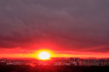Hot red sun behind the silhouettes of city buildings. Summer sunset in the sky haze with night houses