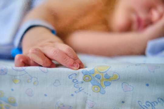 Hand newborn baby with a maternity hospital bracelet on his arm is sleeping in a crib. A newly born child in a clinic bed behind a transparent glass