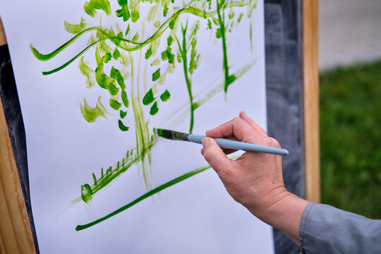 Happy woman draws on the easel with a brush and paints. Woman artist draws nature and trees on paper by the water on the river bank