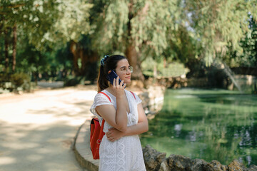 Long shot of a young woman with a white dress and a red backpack talking on the phone in a park