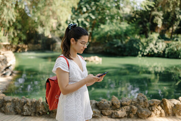Young woman with a white dress and a red backpack using her phone in a park