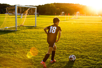 Young boy wearing jersey kicking a soccer ball on field at sunset