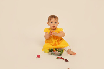 a beautiful little girl in a yellow dress is sitting on a white background next to a small basket with vegetables