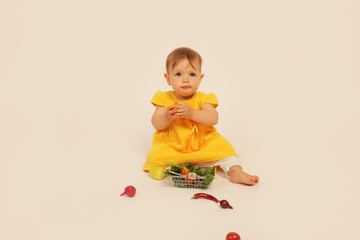 a beautiful little girl in a yellow dress is sitting on a white background next to a small basket with vegetables