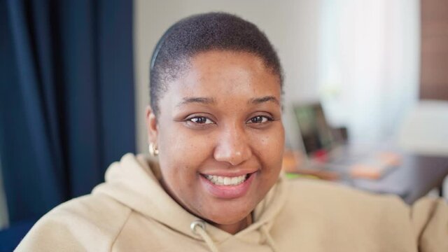 Head and shoulders portrait of beautiful Black plus size young woman in beige hoodie looking at camera with cheese smile. Cheerful plump African female posing at home