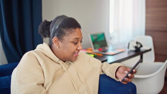 Portrait of chubby Black woman in beige hoodie typing sms on cellphone, looking at camera and smiling. Young female text messaging on phone and posing sitting on sofa while working from home