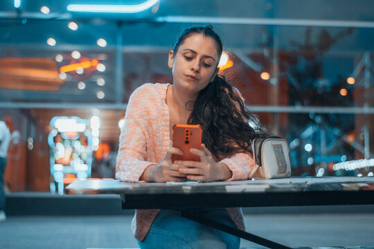 Muchacha joven con seriedad escribiendo por tel&eacute;fono m&oacute;vil esperando en la mesa de un bar para cenar por la noche junto al centro comercial
