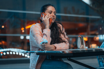 Muchacha joven de cabello rizado llamando por smartphone mientras sonríe contenta en una mesa sentada para ir a la fiesta de la feria con amistades