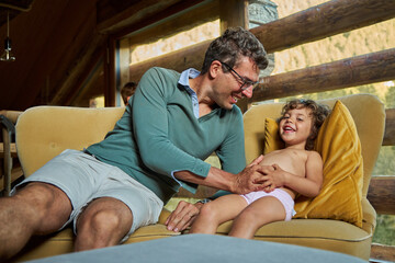 A young curly-haired dad is playing with a little curly-haired girl. Beautiful house made of natural wooden materials. A window with wooden boards. Summer holidays.