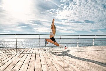 Life balance. Calm and sporty disabled athlete woman in sportswear with prosthetic leg standing in yoga pose on the bridge in front of the sea.