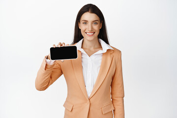 Smiling corporate woman showing smartphone screen flipped horizontal, recommending company website, store online, standing over white background