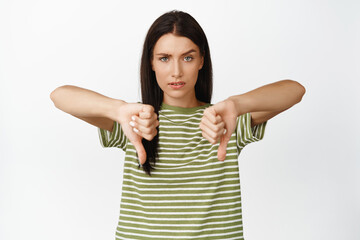 Disappointed brunette woman shows thumbs down, dislike smth, disapprove action, standing over white...