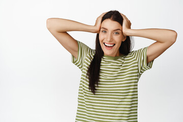 Happy brunette woman looking excited, smiling at camera, holding hands on hair, standing in tshirt over white background