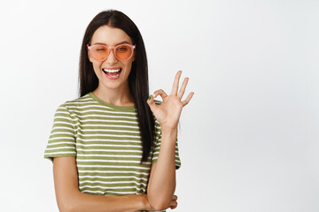 Happy brunette woman in sunglasses, smiling and showing ok sign, concept of summer vacation, white background