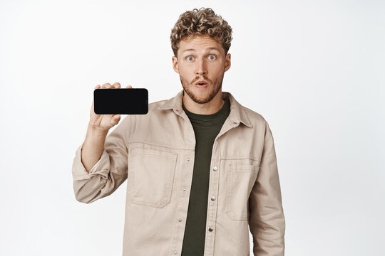 Image of amazed guy showing mobile phone screen horizontally, staring impressed at camera while demonstrating smth cool on smartphone app, white background