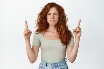 Portrait of moody redhead girl sulking, pointing fingers up at smth unfair, being disappointed,...