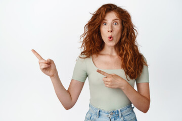 Wow sale there. Amazed redhead girl pointing left, being impressed by sale announcement, showing logo, standing in t-shirt against white background