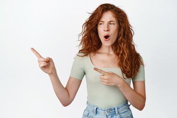 Shocked redhead woman pointing, looking left with offended face expression, standing against white...
