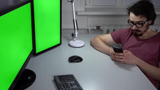 Young man is sitting in office in front of computer with double green screen and using mobile phone. He checks information in smartphone and types something on keyboard. Bearded programmer in glasses.